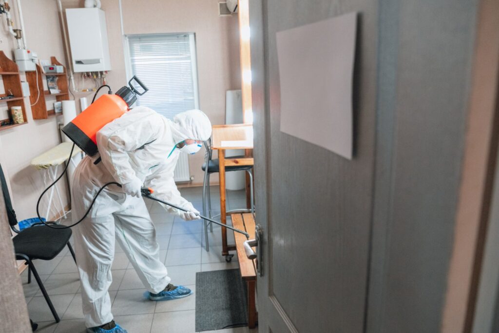 A person in full protective gear uses a sprayer to disinfect a room, focusing on furniture and door areas, in a small, modern indoor space.