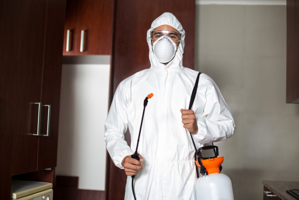 A person wearing a protective white suit, gloves, and a mask stands indoors holding a sprayer, appearing ready to disinfect or sanitize the area.